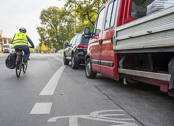Ein Radschutzstreifen wird von Pkws blockiert, während eine Radfahrerin in die Mitte ausweichen muss.