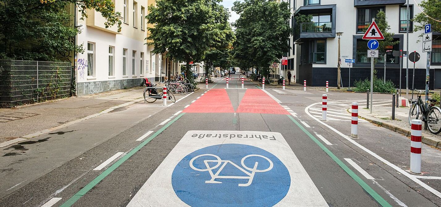 Eine Straße in Berlin: Rechts und links sind Häuserzeilen zu sehen, die Fahrbahn ist mit einem großen Fahrradsymbol versehen. Weiter hinten ist die Fahrbahn rot eingefärbt, Poller dienen als modaler Filter.  